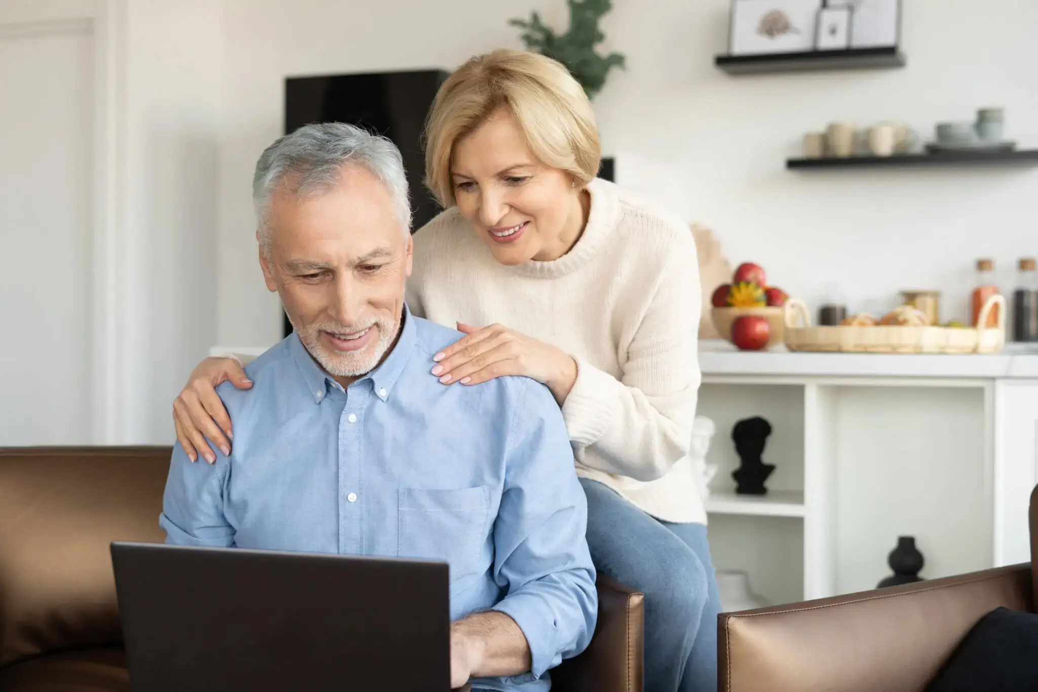 Older woman puts hands on her husband's shoulders encouragingly as he uses his laptop.