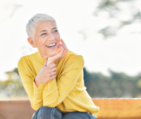 Smiling senior female sitting on a park bench wearing a bright yellow sweater